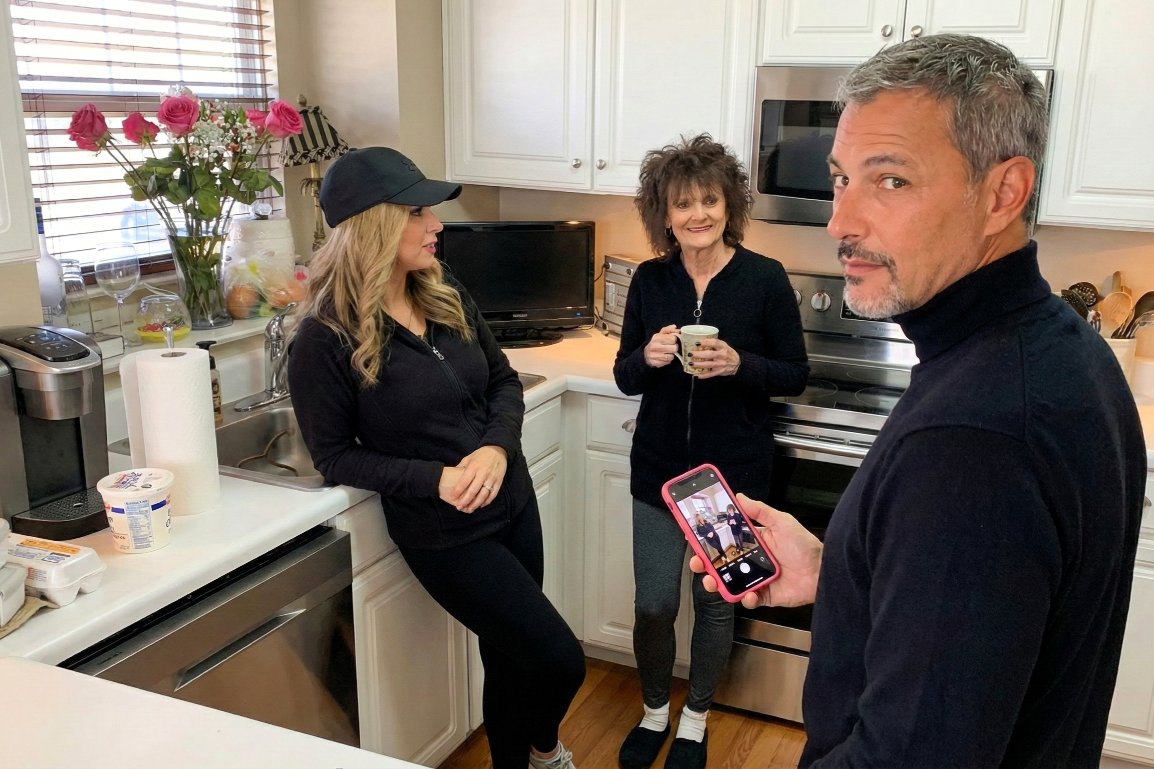 Joey Montes holding a phone, Heather Killebrew in a black cap, and a woman with a mug chatting in the kitchen.