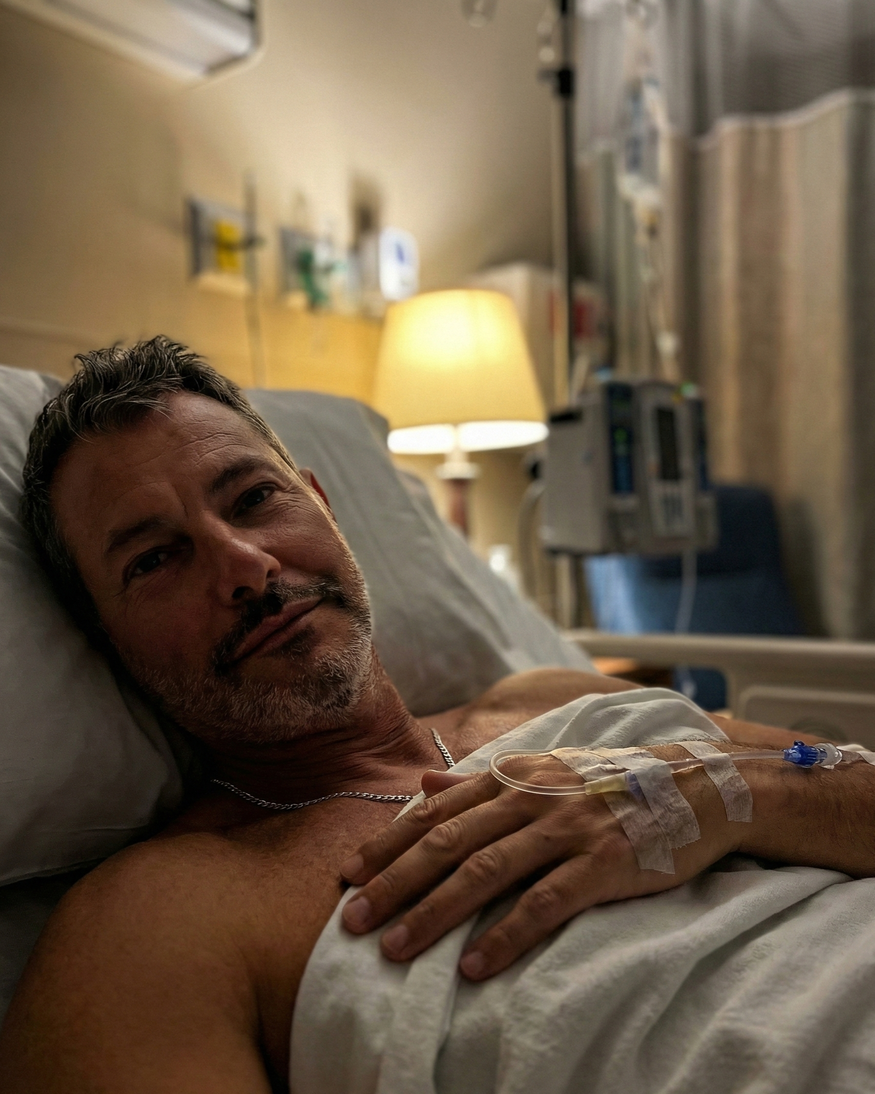 Intimate low-light photo of Joey Montes resting in a hospital bed with an IV in his hand.