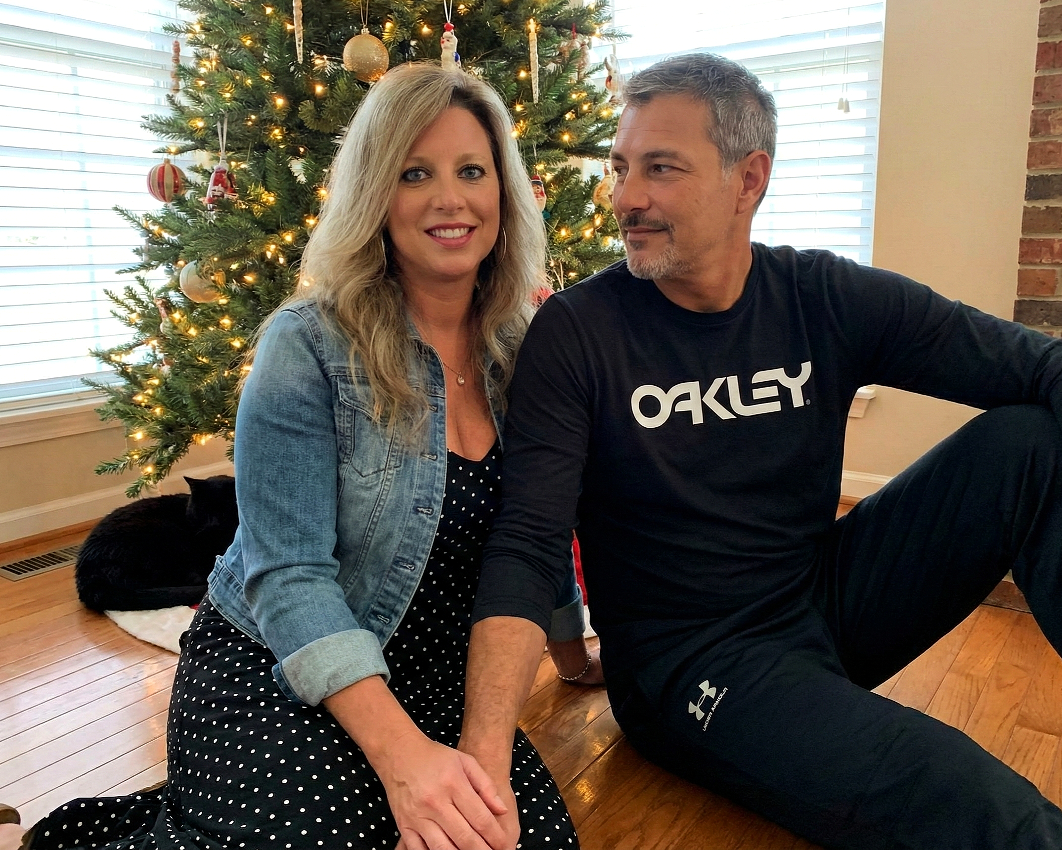 Joey Montes looking at Heather Killebrew while sitting by a Christmas tree with a black dog sleeping in the background.