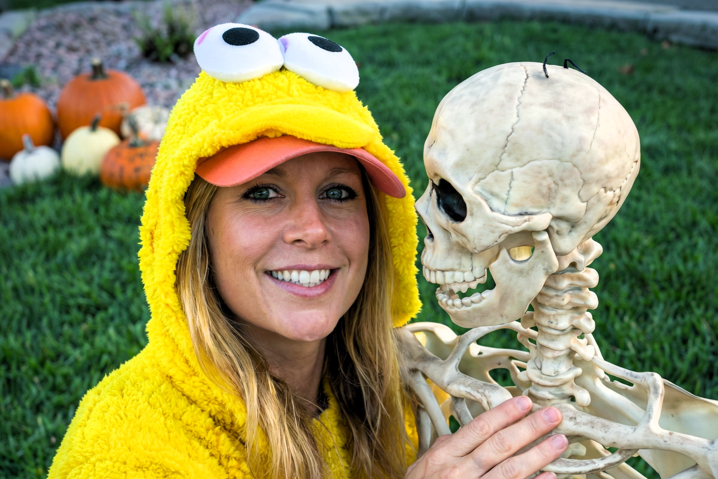 Heather Killebrew in a yellow bird costume smiling next to a plastic skeleton on the grass.