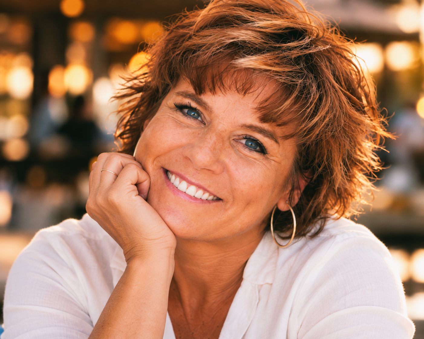 Dawne smiling warmly in June 2016, resting her cheek on her hand, wearing a white blouse and hoop earrings.