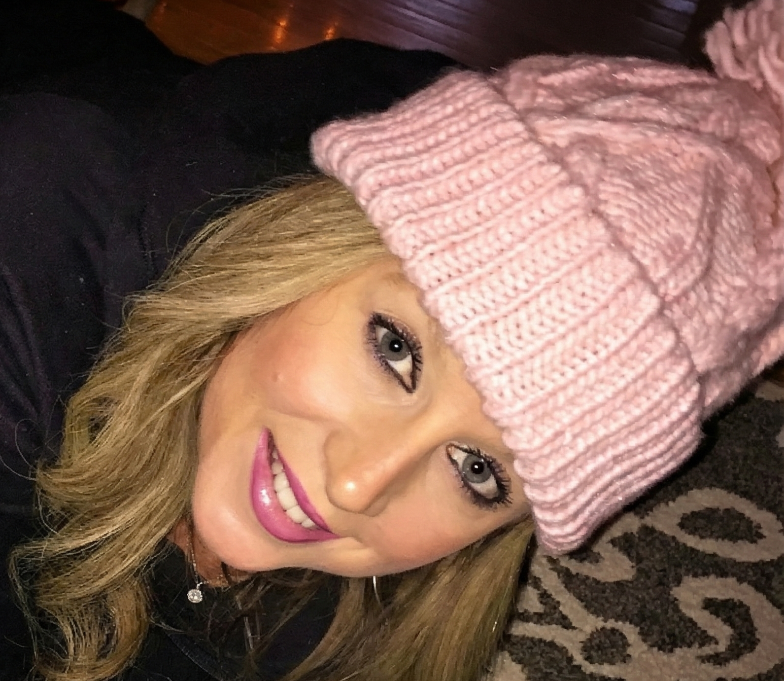 Close-up selfie of Heather Killebrew wearing a pink knit beanie hat and smiling while lying on a patterned rug.