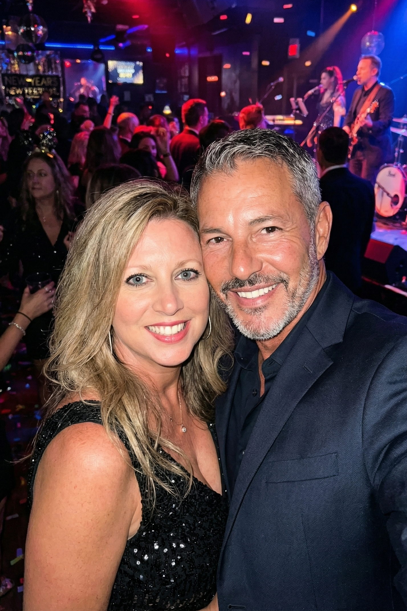 Selfie of Joey Montes and Heather Killebrew smiling at a crowded New Year's party with a live band performing in the background.