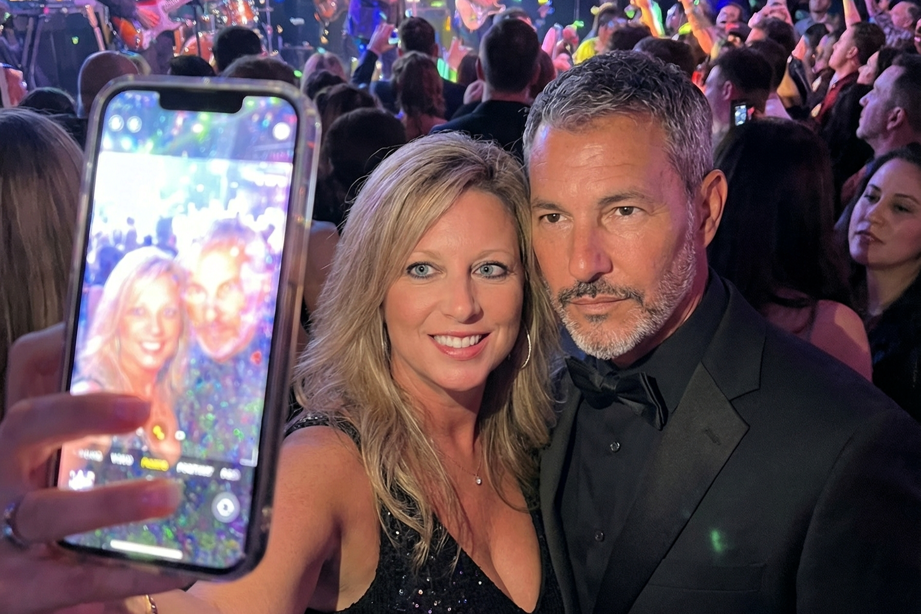 A photo of Heather Killebrew holding up a smartphone to take a selfie with Joey Montes, surrounded by purple party lights and a crowd.