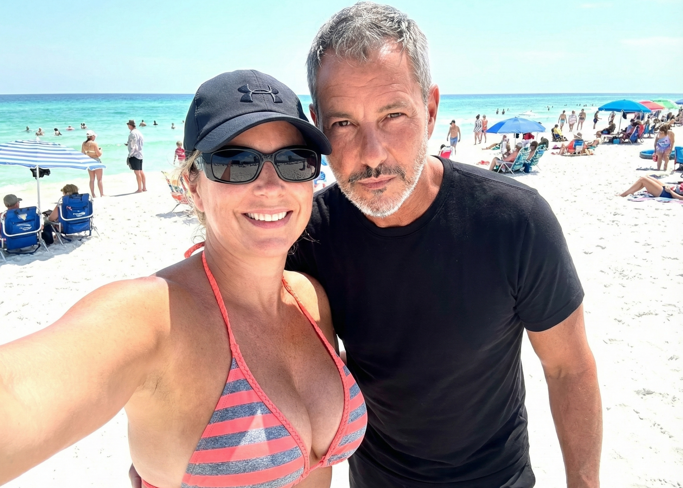 Sunny beach selfie of Joey Montes and Heather Killebrew standing on the sand with blue umbrellas in the background.