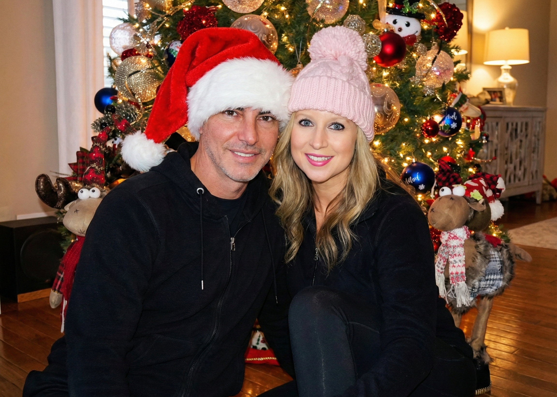 Joey Montes wearing a Santa hat and Heather wearing a pink beanie sitting in front of a Christmas tree.
