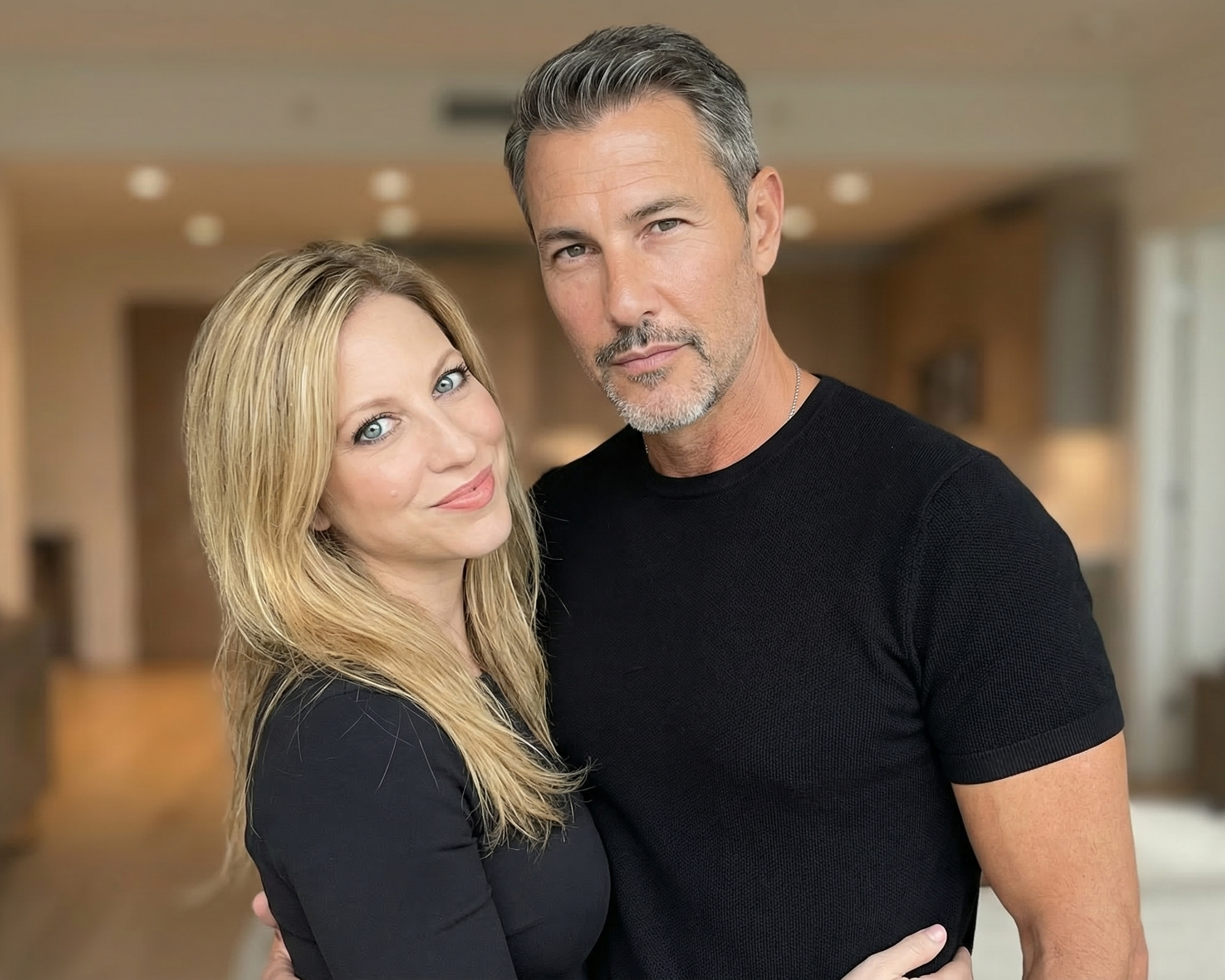 A portrait of Heather smiling and embracing a man in a modern Wildwood home interior, both wearing black.