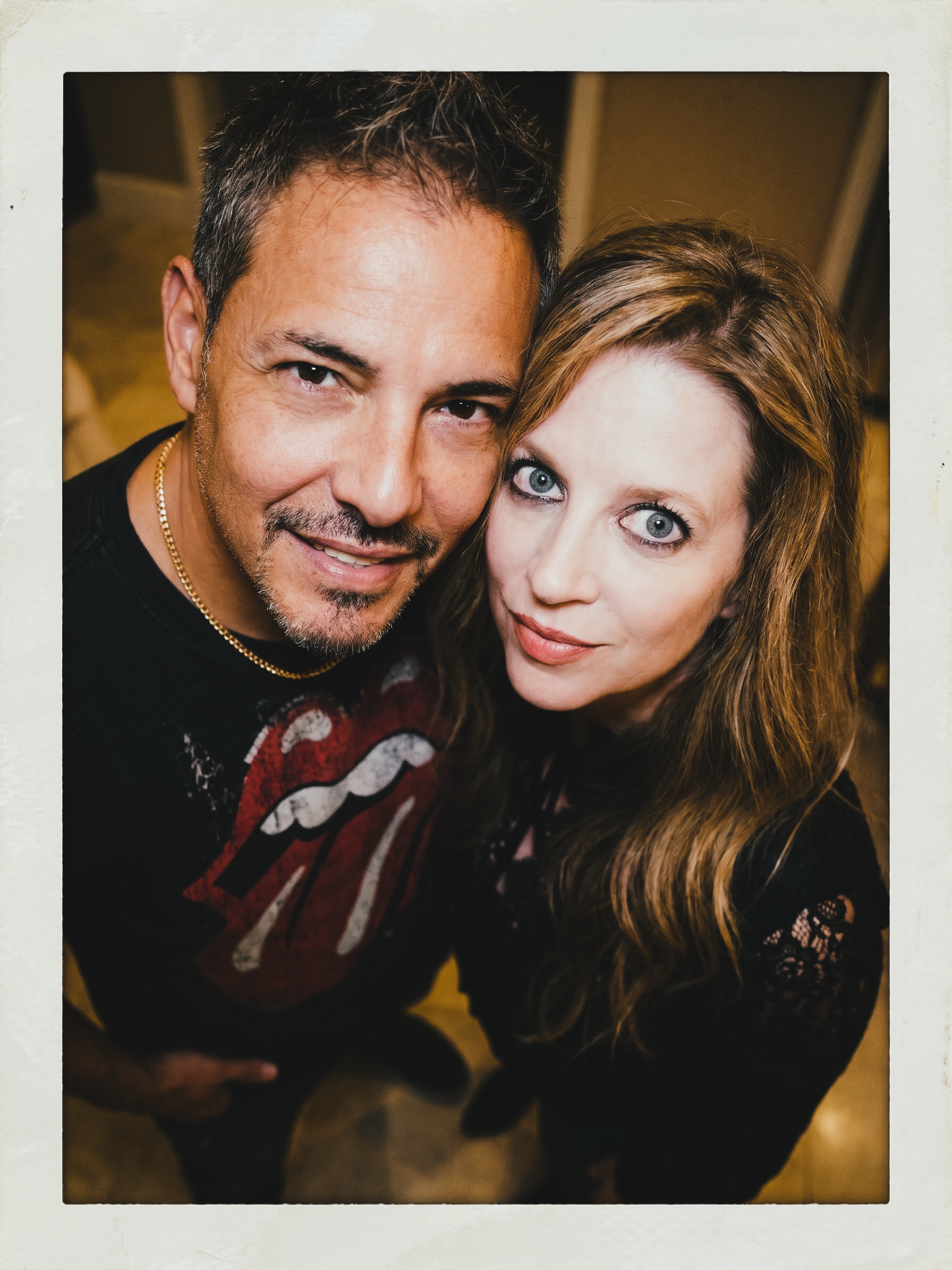 Heather and Joey leaning close together in a warm indoor portrait in Dresden in 2021. Joey wears a black Rolling Stones T-shirt and Heather wears a black lace top. The photograph has a vintage instant-film border.