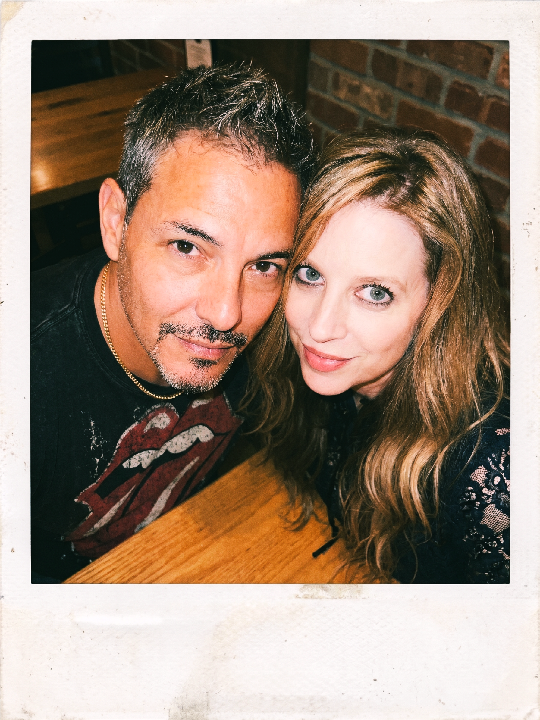 Heather and Joey leaning close together across a wooden table in Dresden in 2021. Joey wears a black Rolling Stones T-shirt and a gold chain. Heather wears a black lace top. The image has a vintage Polaroid-style border.