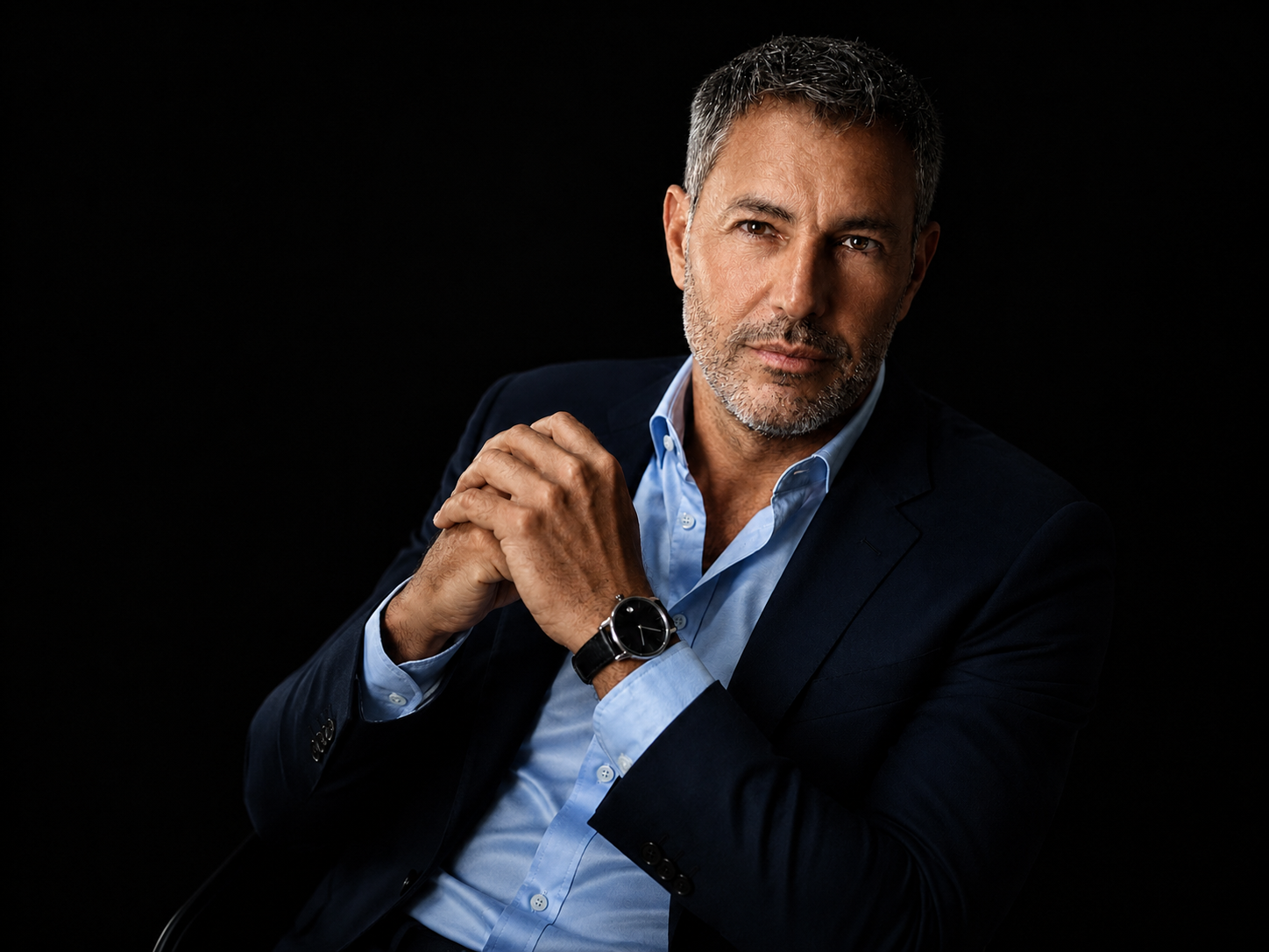 A professional studio portrait of a man in a navy blazer and light blue shirt against a black background.
