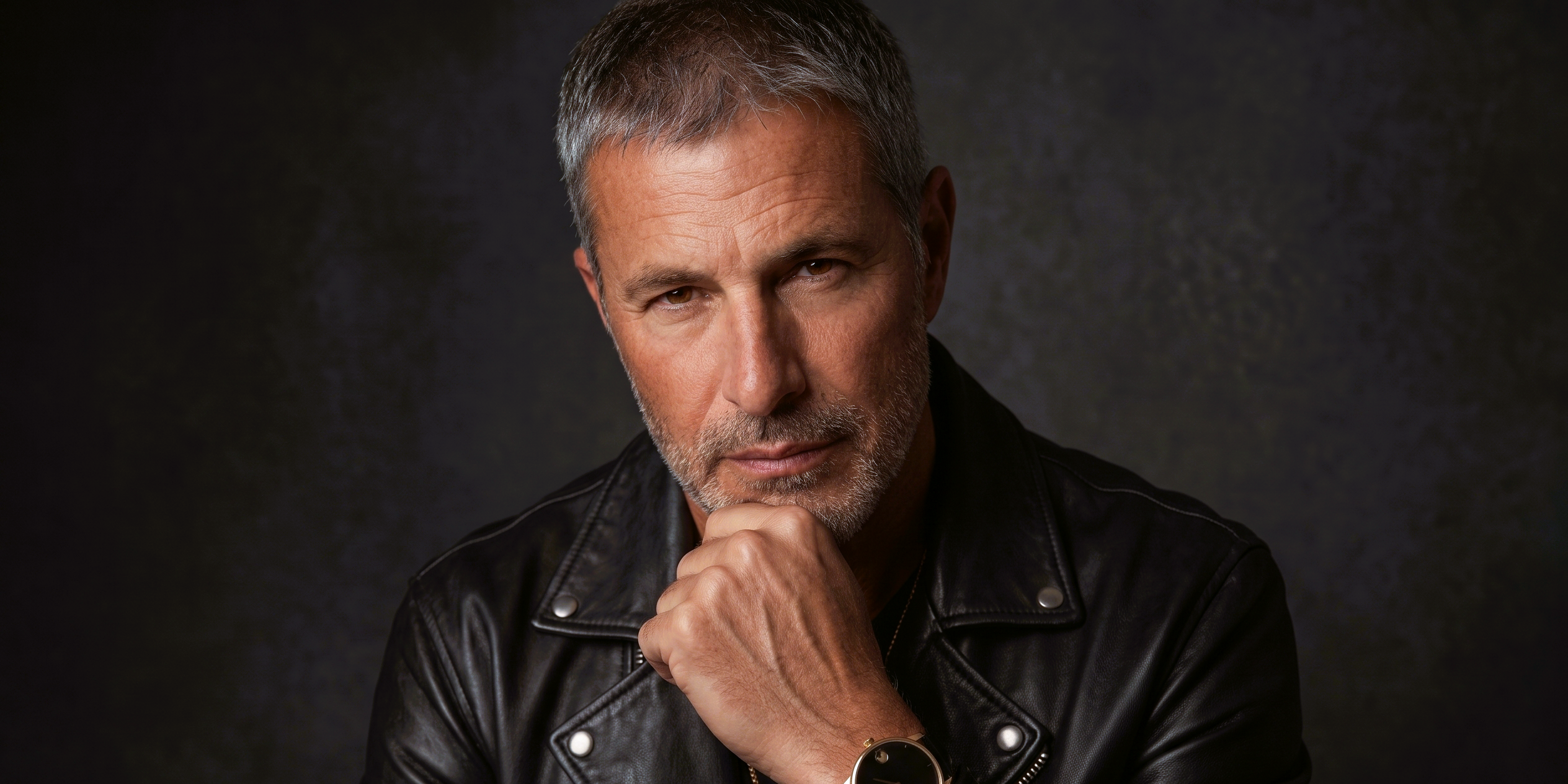 Studio close-up portrait of Joey at age 58 wearing a black leather jacket, resting his chin on his hand with a steady expression.