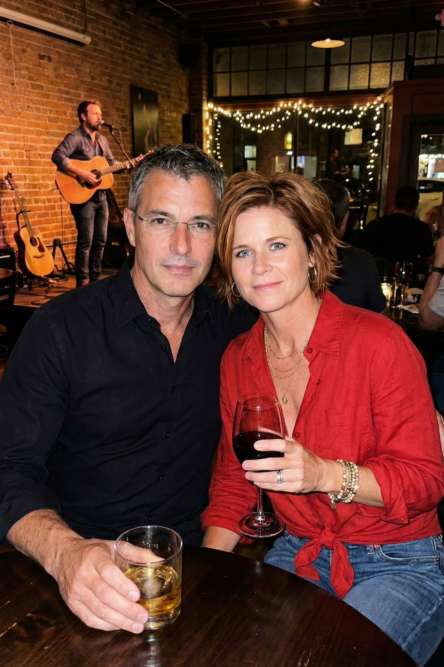 Joey Montes holding a whiskey and Dawne holding a glass of red wine at a table with a musician playing in the background.