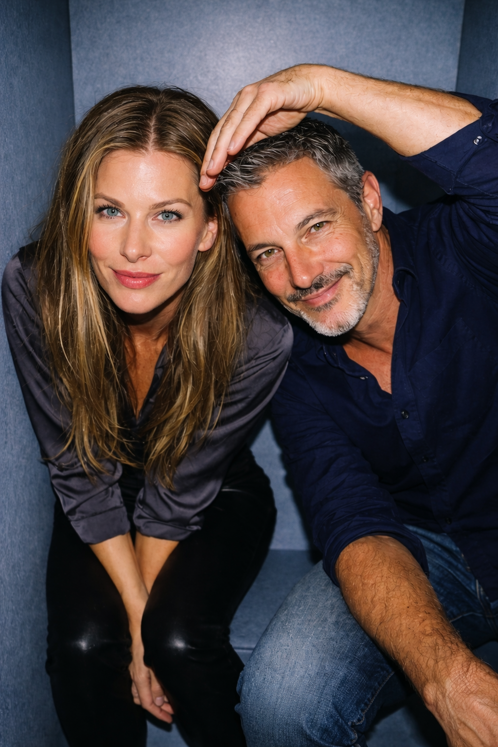 Portrait of a female friend playfully resting her hand on Joey Montes's head, both smiling warmly in a blue studio.