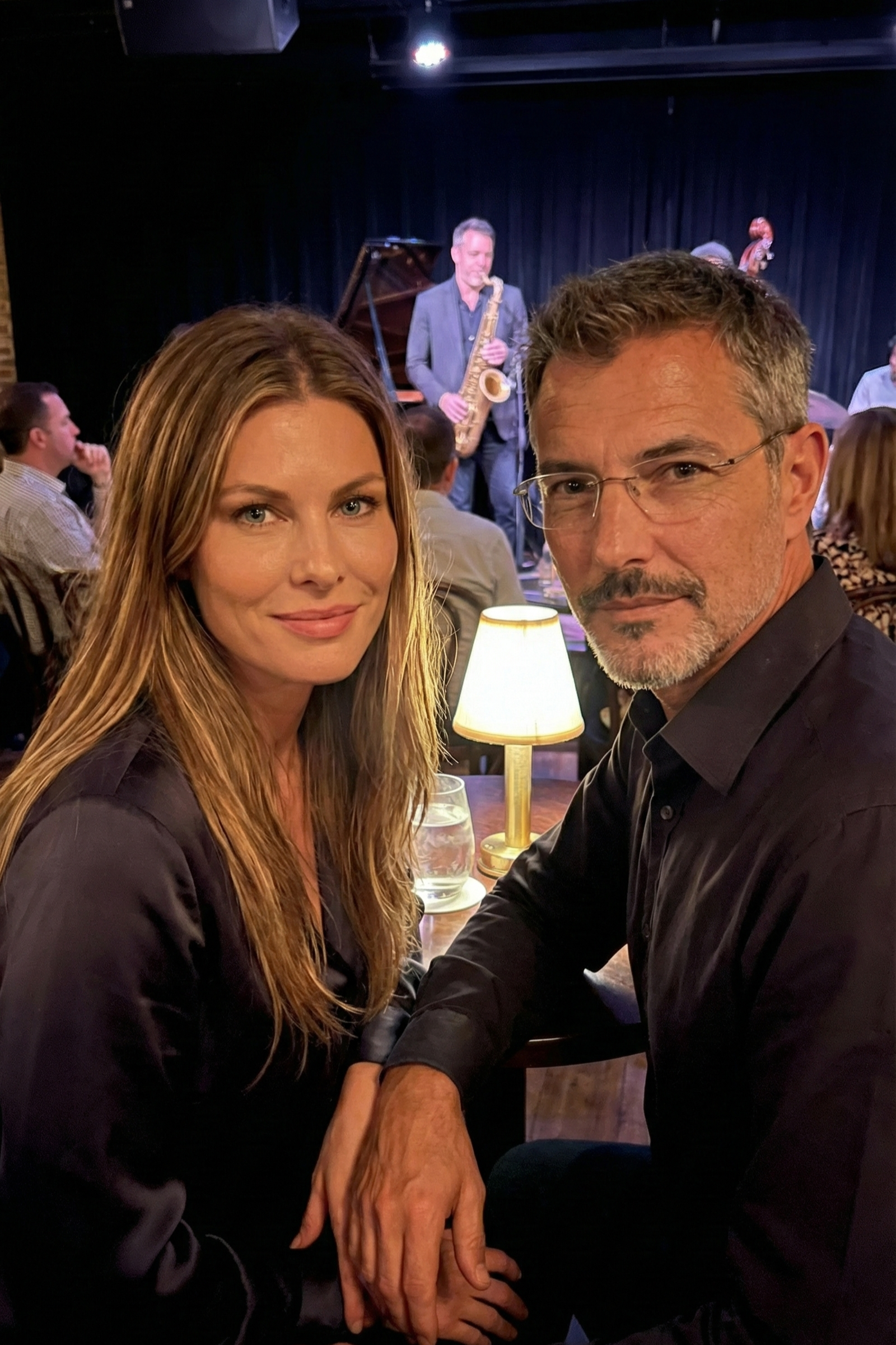 Joey Montes and a friend seated at a jazz club table with a saxophone player performing in the background.