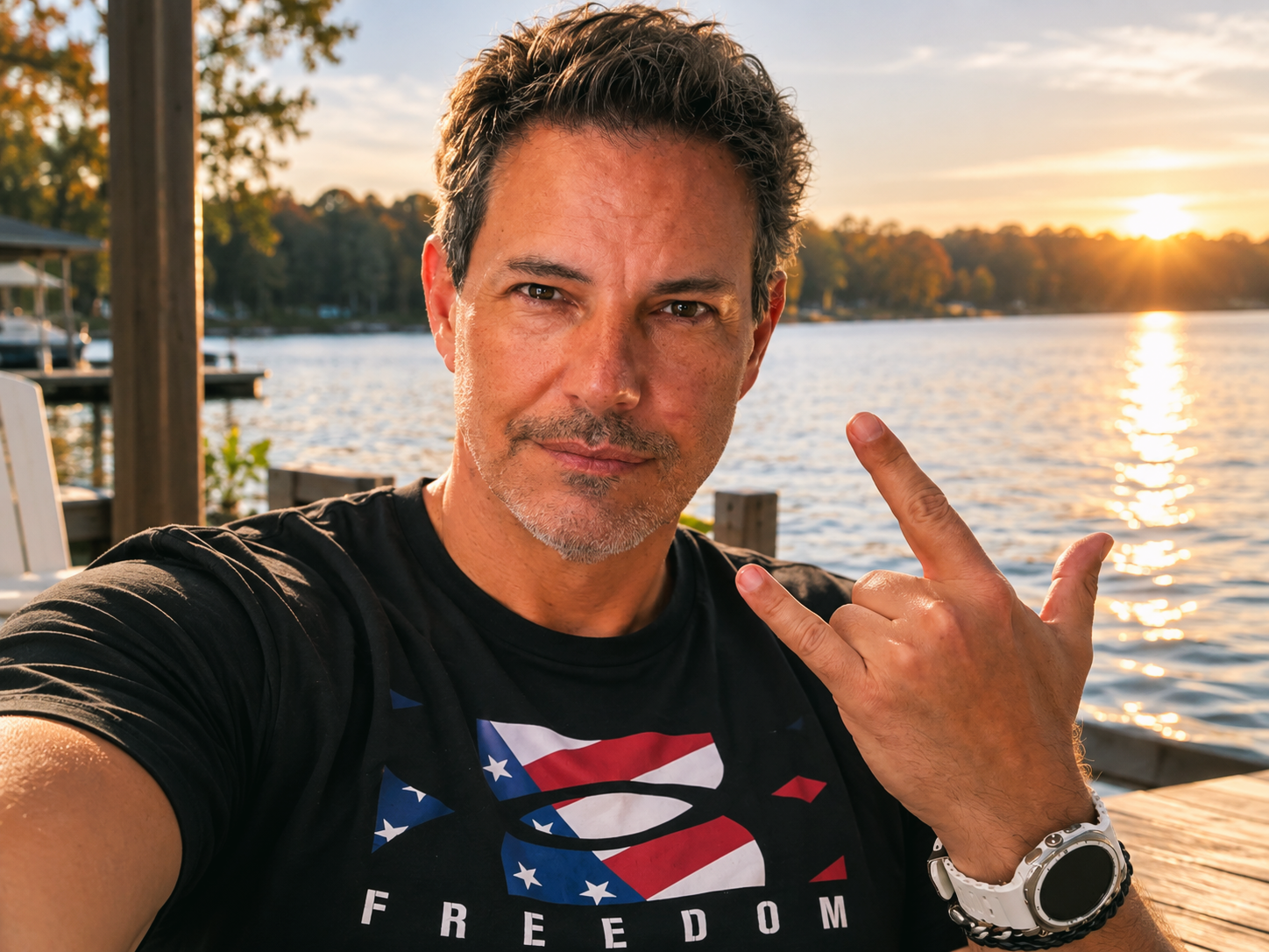 Joey taking a selfie beside the lake at sunset in August 2026, wearing a black Freedom T-shirt and white watch while making a rock and roll hand gesture.